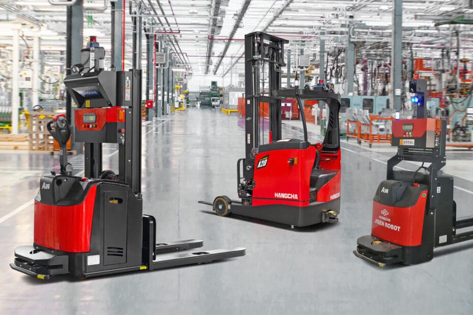 Three red automated guided vehicles (AGVs) positioned on a polished factory floor inside a large industrial facility. Each AGV is equipped with lifting mechanisms for pallet handling and features sensors for navigation. The background shows rows of machinery, safety barriers and overhead lighting in a spacious production environment.